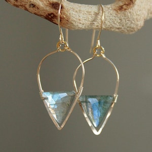 May include: A pair of gold-colored hoop earrings with a teardrop-shaped design. Each earring features a triangular, iridescent gemstone in shades of blue and green, secured with wire wrapping. The earrings are suspended from a curved gold wire.