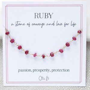 May include: A delicate gold chain necklace featuring small, faceted, deep red ruby beads. The necklace is displayed on a white card with the text RUBY, a stone of courage and love for life, passion, prosperity, protection, and the brand name Otis B.