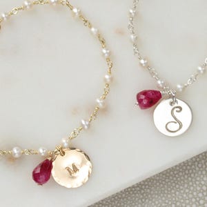 Peut inclure: Deux bracelets délicats avec des perles et une chaîne en or ou en argent. Chaque bracelet comporte un charm rond avec une initiale et une pierre précieuse rouge. Un charm porte la lettre "M", l'autre la lettre "S".