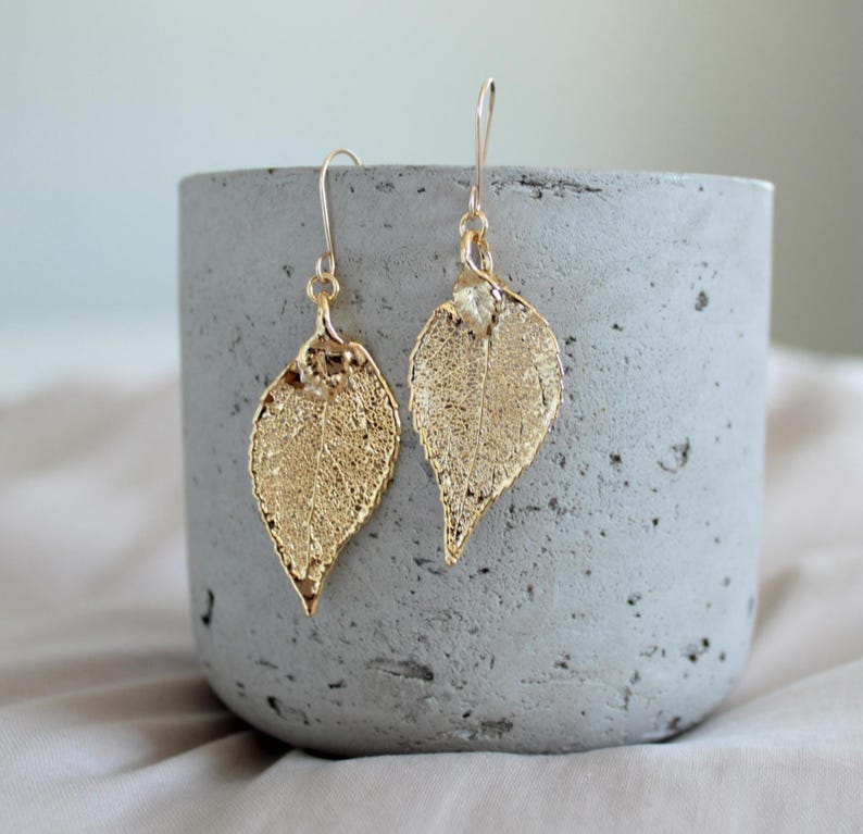 May include: A pair of gold-colored leaf earrings with delicate veining. The earrings are suspended from gold-colored ear wires. The earrings are displayed on a gray concrete container.
