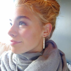 May include: A woman with blonde hair wearing a gray scarf and long, thin gold earrings. The earrings are made of small, rectangular pieces of metal.