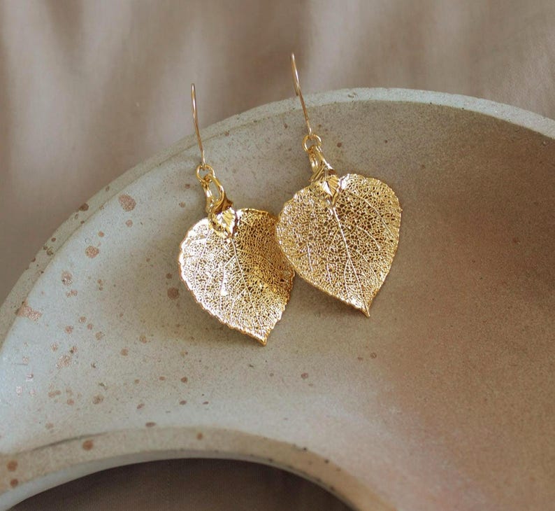 May include: A pair of gold-colored leaf-shaped earrings. The earrings are made of a delicate, lacy material, with visible veins and intricate details. The earrings are suspended from gold-colored hooks. The earrings are resting on a light-colored, textured surface.
