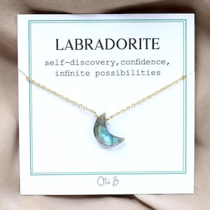 May include: A gold-colored chain necklace with a crescent moon pendant made of labradorite. The pendant is a mix of blue, green, and gray. The necklace is displayed on a white card with the word "LABRADORITE" and other text.