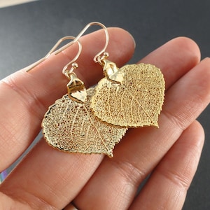 May include: Gold-colored leaf earrings with intricate vein detailing. The earrings feature a delicate, openwork design in the shape of a heart. The earrings hang from gold-colored ear wires. These earrings are a nature-inspired accessory.