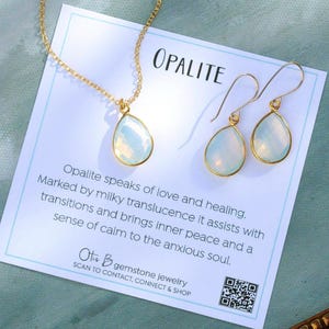 May include: A gold-toned necklace and earrings set featuring opalite teardrop pendants. The jewelry is displayed on a card with the word "OPALITE" and text about the gemstone's properties. The earrings have a hook closure.