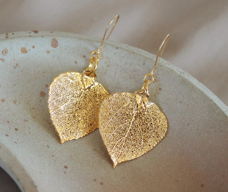 May include: A pair of gold-colored leaf earrings. The earrings are shaped like leaves with intricate vein detailing. They hang from delicate gold-colored ear wires. The earrings are displayed on a light gray surface.
