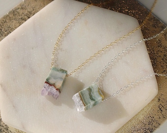 May include: Two necklaces with green and purple agate stone pendants. The necklaces are on a white surface. One necklace has a gold chain and the other has a silver chain.
