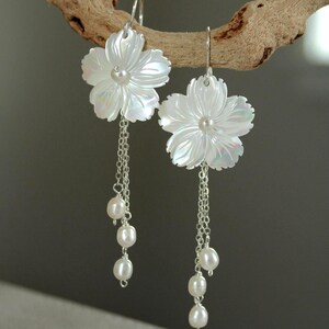 May include: White flower-shaped earrings with a pearl center and dangling pearl accents. The earrings are suspended from silver hooks and chains. The floral design has iridescent qualities, and the pearls are white.