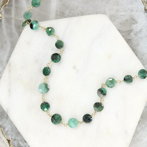 May include: A delicate necklace featuring small, round, faceted green emerald beads linked together by a thin gold-coloured metal chain. The beads vary in shade from light to dark green and are arranged in a continuous, flowing, curved line pattern.