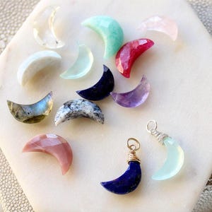 May include: A collection of crescent moon-shaped gemstones in various colors including clear, light blue, pink, dark blue, purple, and iridescent gray. Two of the moon-shaped stones are fitted with silver-toned wire loops for use as pendants.