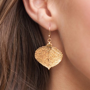 May include: Gold-colored leaf-shaped earrings. The earrings are made of a delicate, lacy material, with a hook closure. The leaf design is detailed with visible veins, creating a natural and elegant look. The earrings are shown worn on an ear.