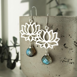 May include: Silver lotus flower earrings with faceted blue and brown gemstone drops. The earrings are hanging against a gray concrete background. The lotus flower design is open and airy.