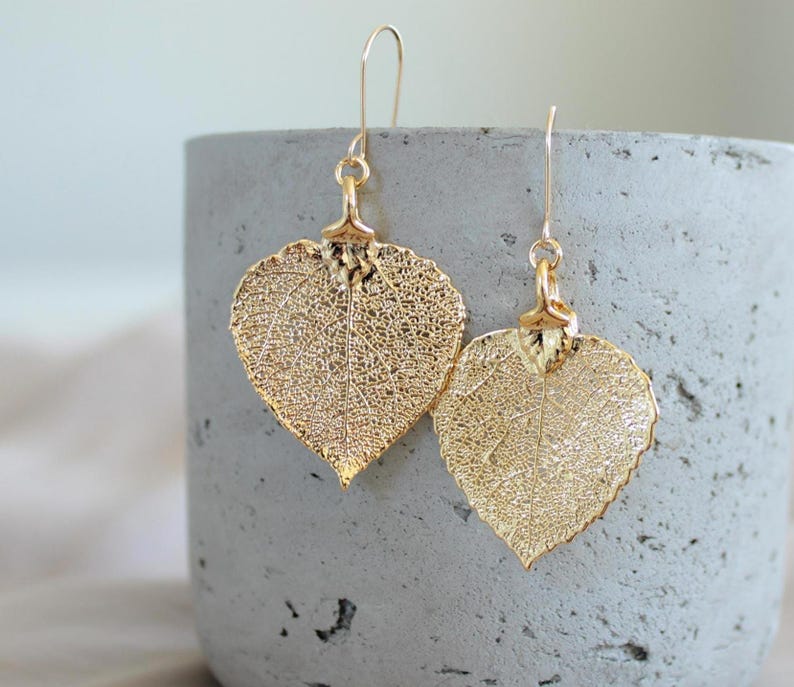 May include: Gold-colored leaf-shaped earrings with intricate vein detailing. The earrings hang from delicate gold-tone ear wires. The leaves are heart-shaped and have a textured appearance, creating a natural and elegant look. The earrings are displayed against a gray background.