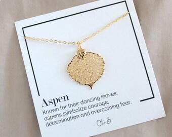 May include: A gold-colored necklace with a leaf pendant. The pendant is a detailed replica of a leaf. The necklace is displayed on a white card with the word "Aspen" and text about the symbolism of aspens.