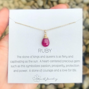 Könnte beinhalten: Eine goldene Kettenkette mit einem Rubin-Anhänger. Der Anhänger ist mit Golddraht umwickelt. Die Karte lautet "RUBY Der Stein der Könige und Königinnen ist so feurig und fesselnd wie die Sonne. Ein herzhaftes, wertvolles Juwel wie dieses symbolisiert Leidenschaft, Wohlstand, Schutz und Kraft. Ein Stein des Mutes und der Liebe zum Leben. Otis & Jewelry"