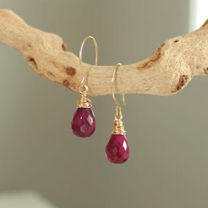 May include: A pair of drop earrings featuring faceted, teardrop-shaped deep pink gemstones. Each stone is securely wrapped in thin gold-toned wire at the top and suspended from a matching gold-toned curved ear wire hook for a simple, elegant look.