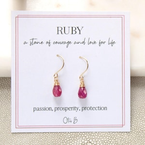 May include: A pair of ruby earrings with gold-colored wire wrapping and hooks. The earrings hang in front of a white card with the word "RUBY" and the text "a stone of courage and love for life" and "passion, prosperity, protection".