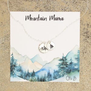 May include: A silver necklace with a mountain and a pine tree charm. The necklace is on a white card with the words "Mountain Mama" and "Otis B" printed on it. The card has a watercolor background of mountains and trees.