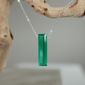 May include: A silver necklace with a rectangular, emerald-green pendant. The pendant is a long, faceted gemstone suspended from a delicate chain. The background is a neutral gray, with a piece of driftwood in the upper left corner.