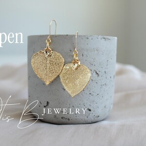 May include: Gold-colored leaf-shaped earrings with a delicate, textured design. The earrings hang from gold-colored hooks. The word "Aspen" is written in a script font. The earrings are displayed on a gray concrete cylinder. The words "Otis B Jewelry" are written in a script font.