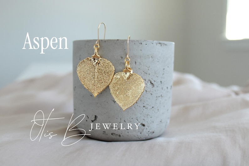 May include: Gold-colored leaf-shaped earrings with a delicate, textured design. The earrings hang from gold-colored hooks. The word "Aspen" is written in a script font. The earrings are displayed on a gray concrete cylinder. The words "Otis B Jewelry" are written in a script font.