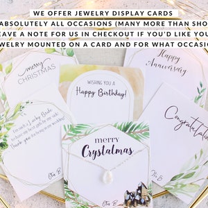 May include: A collection of jewelry display cards for various occasions, including Christmas, birthdays, and anniversaries. The cards feature watercolor designs and text such as "Happy Anniversary" and "Merry Christmas". The cards are displayed with a necklace.