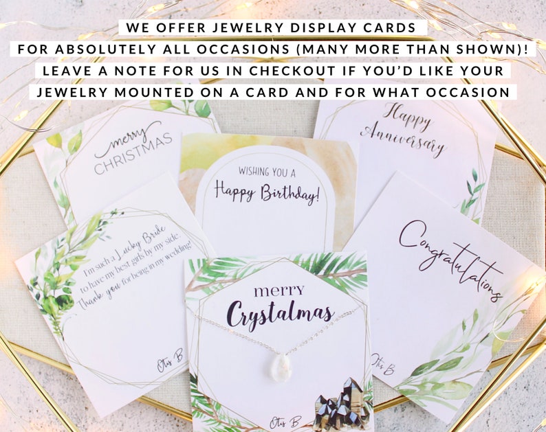 May include: A collection of jewelry display cards for various occasions, including Christmas, birthdays, and anniversaries. The cards feature watercolor designs and text such as "Happy Anniversary" and "Merry Christmas". The cards are displayed with a necklace.