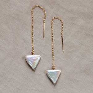 May include: A pair of gold-toned chain earrings with a triangular, iridescent pearl pendant. The earrings have a delicate chain that hangs down, with a straight bar at the top for insertion into the ear. The background is a solid, neutral color.