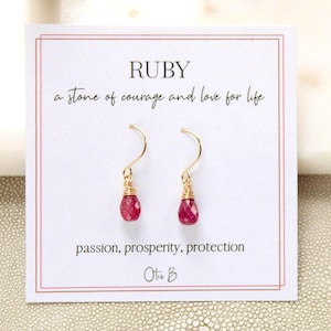 May include: A pair of drop earrings featuring faceted, teardrop-shaped ruby stones suspended from gold-toned wire hooks. The earrings are displayed on a white card printed with the text RUBY, a stone of courage and love for life, passion, prosperity, protection, and Otis B.