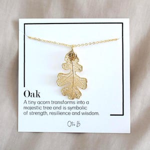May include: Gold-colored oak leaf pendant necklace. The leaf design is intricate and detailed, suspended from a delicate chain. The card behind the necklace reads "Oak" and describes the oak tree's symbolism of strength and wisdom.