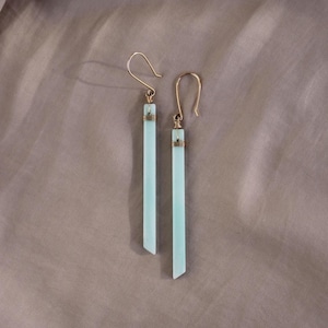 May include: A pair of long, light blue earrings with gold wire accents. The earrings are made of a smooth, polished material and have a slightly pointed shape.