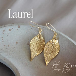 May include: Gold-colored leaf earrings with a delicate, veined design. The earrings are displayed on a speckled, light gray surface. The word "Laurel" is above the earrings, and the text "Otis B Jewelry" is below.