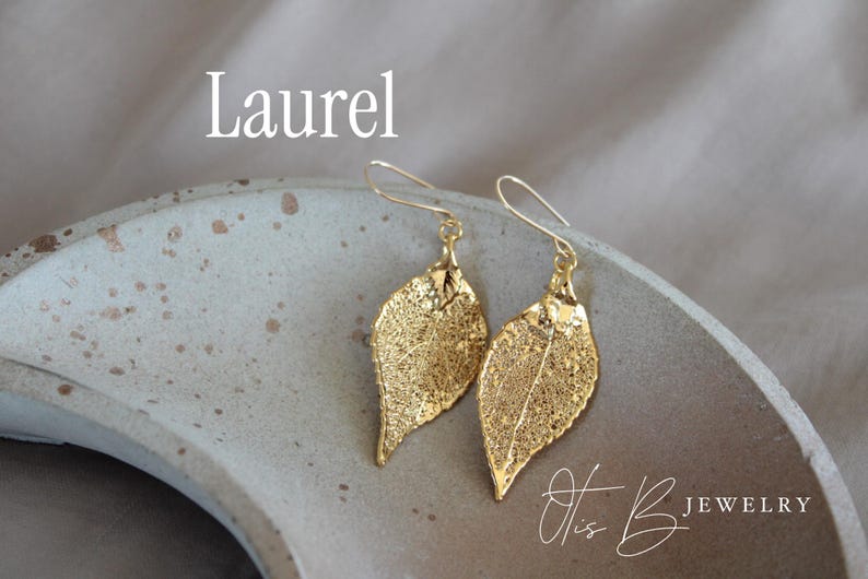 May include: Gold-colored leaf earrings with a delicate, veined design. The earrings are displayed on a speckled, light gray surface. The word "Laurel" is above the earrings, and the text "Otis B Jewelry" is below.