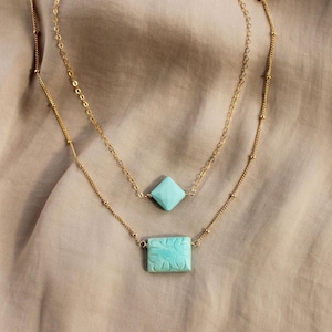 May include: Two gold-colored necklaces with turquoise pendants. The top necklace features a diamond-shaped pendant, while the bottom necklace has a rectangular pendant with a floral design. The necklaces are displayed on a neutral-toned fabric.
