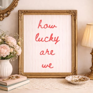 May include: A framed print with the words "how lucky are we" in red script. The frame is gold and ornate, and the background is a striped pattern. The print is displayed on a white table with a vase of flowers and a lamp.