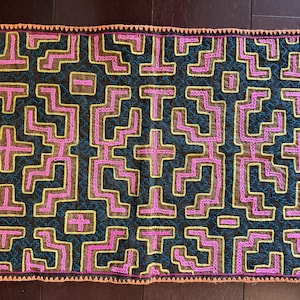 May include: A rectangular textile piece featuring a dark background with intricate geometric embroidery in bright pink, yellow, and teal thread. The edges are finished with a vibrant orange crocheted border, creating a bold contrast.