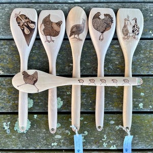 May include: Six wooden spoons with different designs of chickens and birds burned into the wood. The spoons are hanging on a wooden surface.