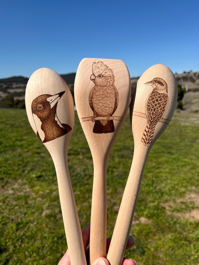 May include: Three wooden spoons with different Australian birds burned into the surface. The spoons are held in a hand with the tips of the spoons pointing towards the camera.