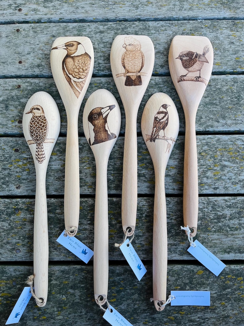 May include: Six wooden spoons with different Australian bird designs burned into the wood. The spoons are arranged in a row on a wooden surface. The birds include a kookaburra, a cockatoo, a wren, a magpie, and two different types of black and white birds.