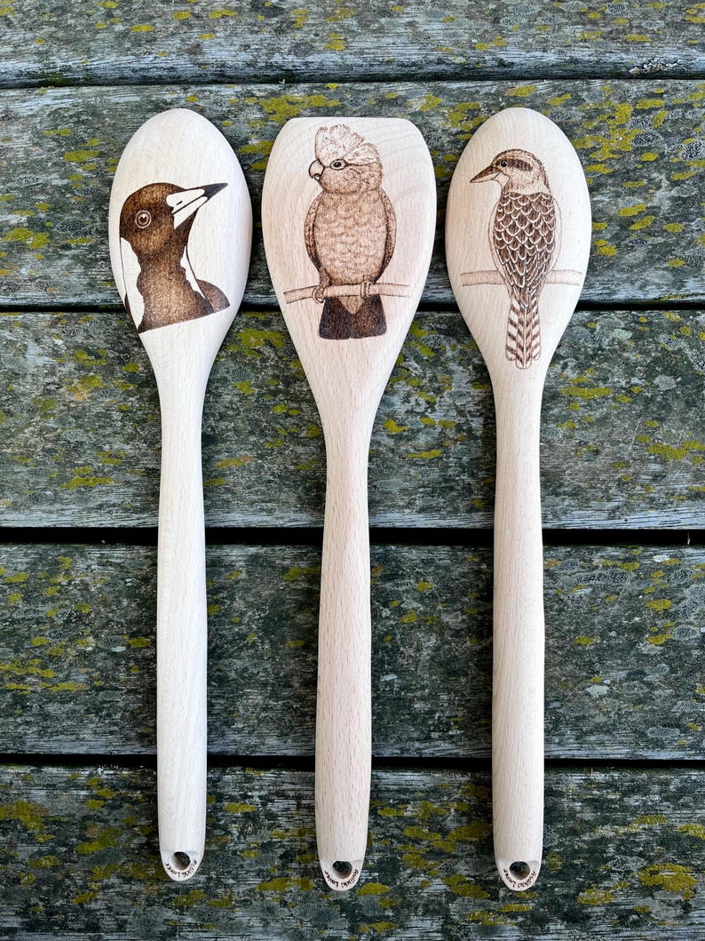 May include: Three wooden spoons with different Australian birds burned into the front of each spoon. The spoons are all natural wood color.