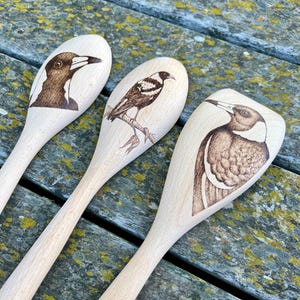 May include: Three wooden spoons with black and white bird illustrations burned into the surface. The birds are Australian Magpies.