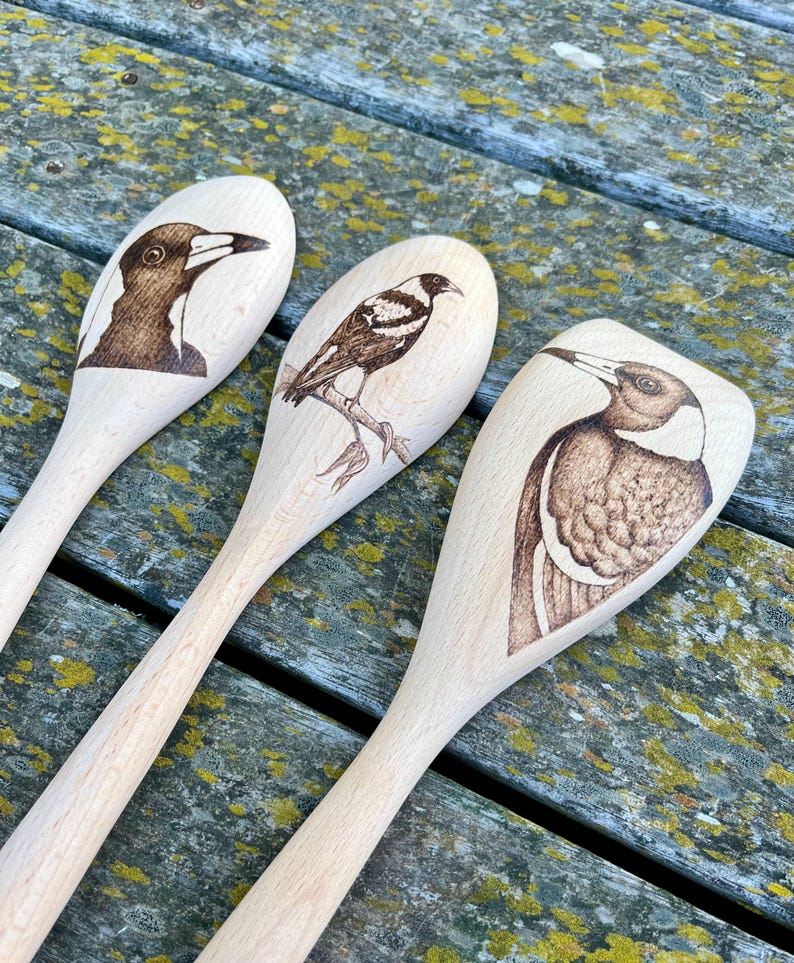 May include: Three wooden spoons with black and white bird illustrations burned into the surface. The birds are Australian Magpies.