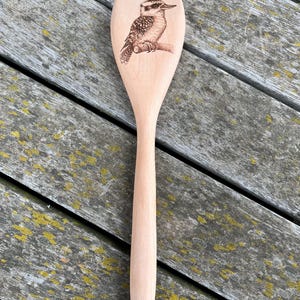 May include: A wooden spoon with a laser-etched design of a kookaburra bird. The spoon is light brown and has a hole at the end of the handle.