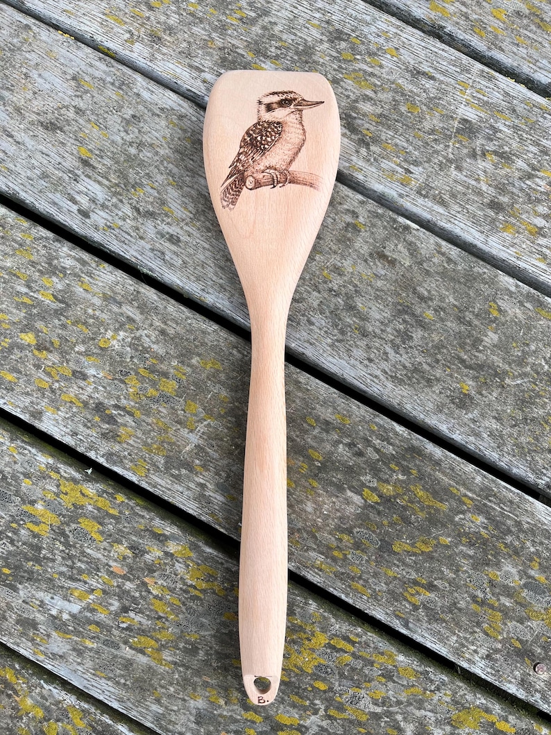 May include: A wooden spoon with a laser-etched design of a kookaburra bird. The spoon is light brown and has a hole at the end of the handle.