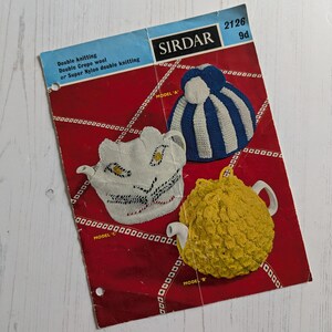 May include: Vintage Sirdar knitting pattern featuring three tea cozy designs. The pattern includes a blue and white striped cozy, a white cozy with black and yellow accents, and a yellow cozy. The pattern is on a red background.