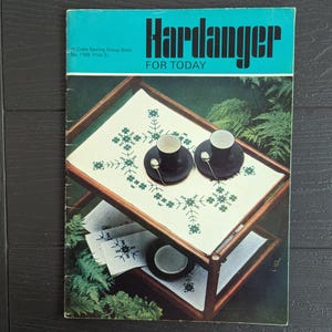 May include: A vintage instructional booklet titled Hardanger FOR TODAY, featuring a wooden serving cart with a white cloth embroidered with green geometric patterns, two black cups with spoons, and additional folded embroidered linens.