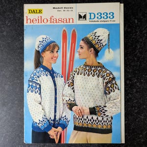 May include: A vintage knitting pattern booklet titled DALE heilo fasan Modell Davos D333. The cover features two people wearing white sweaters and matching hats with blue and gold geometric patterns, standing in front of a pair of red skis.