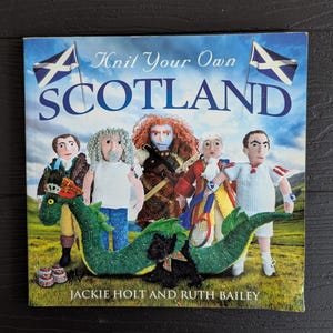 May include: A softcover book titled Knit Your Own SCOTLAND by Jackie Holt and Ruth Bailey. The cover features knitted figures including a green sea monster, a black terrier, and five human characters against a Scottish landscape with two flags.