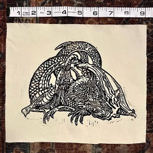 May include: A black and white linocut print of a dragon on cream-colored paper. The dragon is depicted with intricate scales and details, with its body curled and wings spread. The print is approximately 4 inches wide.