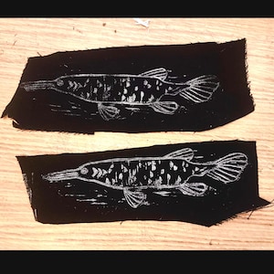 May include: Two black fabric patches with white outlines of a fish with a long snout. The fish is a gar, a type of freshwater fish.